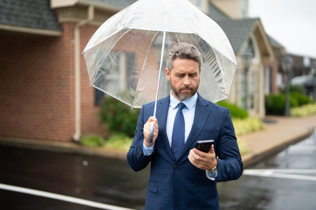 Businessman talks on the phone while standing in the rain. Man in suit with umbrella and makes a mobile phone call. Business man checks his smartphone on a rainy street. Call while rain drops fall.の写真素材
