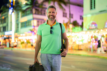 Hispanic tourist traveler on Miami vibrant streets at night. A tourist with a suitcase in a tropical cityscape. Summer tourism in USA. Middle-aged tourist in the night lights of Miami. Mature tourist.の写真素材