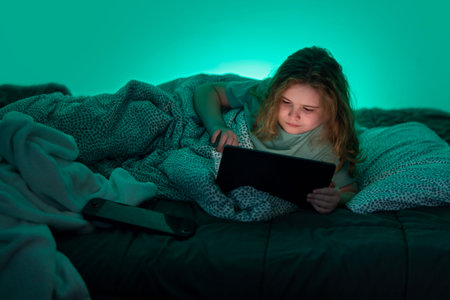 Teenager using tablet under blanket in bed at night. Child lying in bed with tablet at night. Kid...