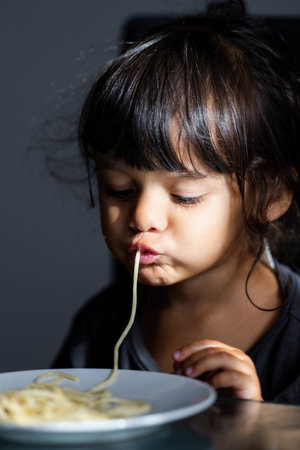 Funny messy face with spaghetti. Toddler eating noodles. Cute childhood emotion during eating. Kid eating spaghetti meal at home. Messy spaghetti mouth and lips moment. Toddler eating pasta indoors.の写真素材