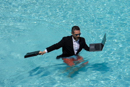 Business man in suit resting in swimming pool. Summer freelancer working in pool. Funny businessman with laptop on sea beach. Guy enjoying travel and beach. Creative online strategy.の写真素材