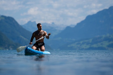 Paddleboarding athlete on calm lake. Paddleboarding man during summer Alps workout. Muscular man on...