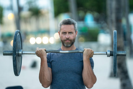 Fit man lifting barbell outdoor. Weight training with barbell in public park. Strong arms and shoulders lifting barbell. Sport model doing barbell. Athletic lifestyle with outdoor barbells exercises.
