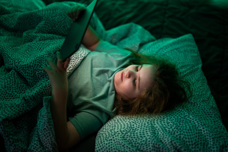Kid watching online videos on tablet in bed at night. Teenager scrolling apps on tablet in bed at night. Child using tablet during bedtime routine in bed at night. Kid under blanket with tablet in bed.