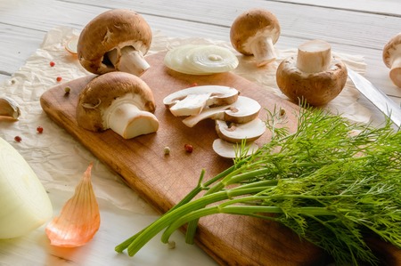 Brown champignon mushrooms on a cutting board. White wooden desk herbs and onions. View angle of 45 degrees.の写真素材