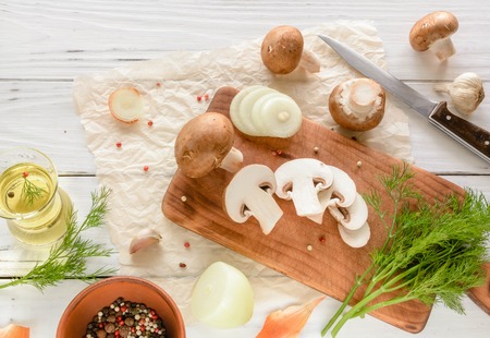 Brown champignon mushrooms on a cutting board. White wooden desk herbs and onions. View angle of 45 degrees.の写真素材