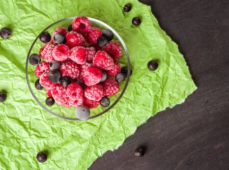 Frozen raspberries in a glass saucer. Frost on the berries. Dark and green background. Green crumpled paper.の写真素材