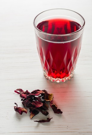 Hibiscus tea in a glass mug on a wooden table among rose petals and dry tea custard. Copy space.の写真素材