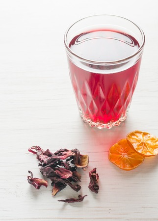 Hibiscus tea in a glass mug on a wooden table among rose petals and dry tea custard. Copy space.の写真素材
