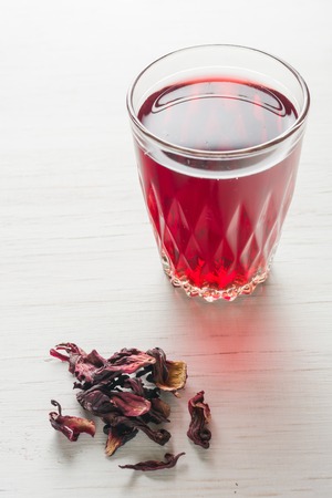 Hibiscus tea in a glass mug on a wooden table among rose petals and dry tea custard. Copy space.の写真素材