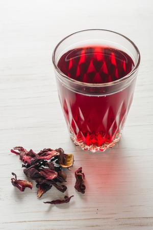Hibiscus tea in a glass mug on a wooden table among rose petals and dry tea custard. Copy space.の写真素材