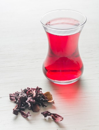 Hibiscus tea in a glass mug on a wooden table among rose petals and dry tea custard. Copy space.の写真素材