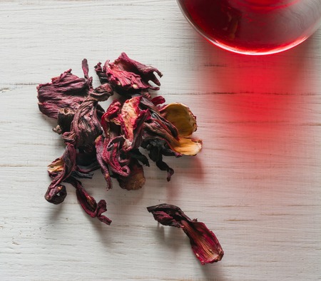 Hibiscus tea in a glass mug on a wooden table among rose petals and dry tea custard. Copy space.の写真素材
