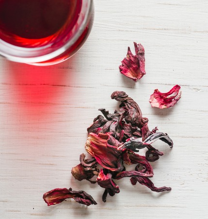 Hibiscus tea in a glass mug on a wooden table among rose petals and dry tea custard. Copy space.の写真素材