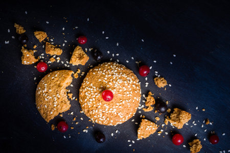Oatmeal cookies are their pieces and crumbs with red berries on a black wooden background. Place for...