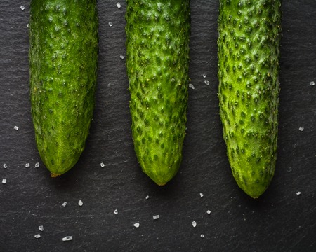 A few fresh green cucumbers lying side by side on a black stone background. A minimalistic photograph. Copy space.の写真素材