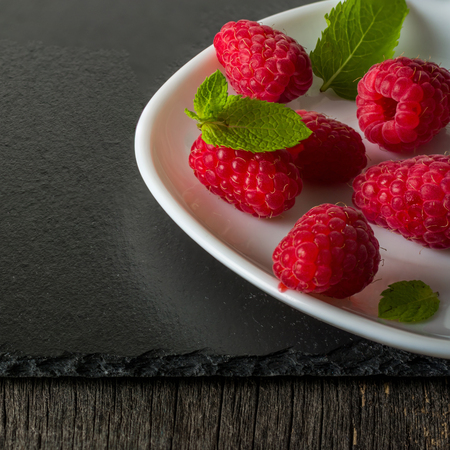 Ripe juicy raspberries on a plate on dark background. Fresh green mint leaves. View from above.の写真素材