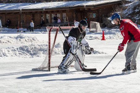 Young boy playing ice hockey in front of goal on outdoor skating rink, with dad trying to score a...