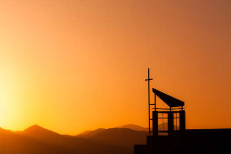 Silhouetted cross against dramatic sunset sky over mountainsの写真素材