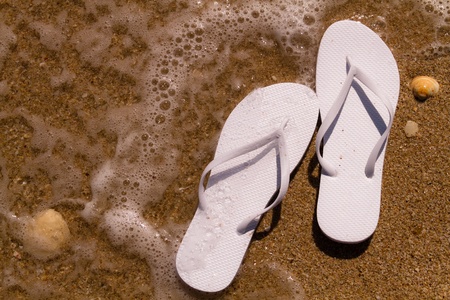 White flip flops laying on the sand with ocean water washing up on shore and sea shellsの写真素材