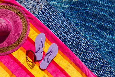 Pair of light purple flip flops by the pool on a bright orange, pink, red and yellow striped towel with sunglasses and big pink floppy hatの写真素材