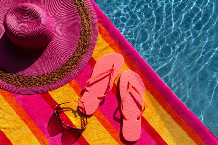 Pair of orange flip flops by the pool on a bright orange, pink, red and yellow striped towel with sunglasses and big pink floppy hatの写真素材