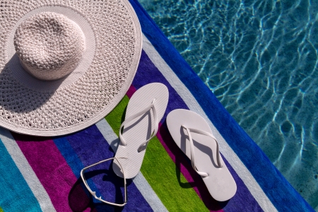 Pair of white flip flops by the pool on a bright blue, green, purple and white striped towel with sunglasses and big white floppy hatの写真素材