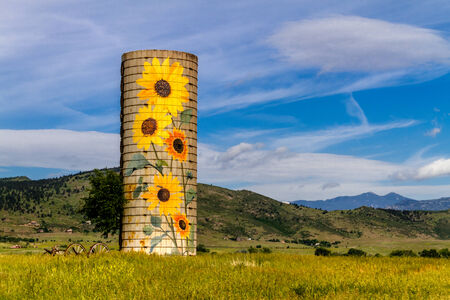 Rural farm silo with painted sunflowers and old farm equipment on sunny summer morning in mountain foothillsのeditorial素材