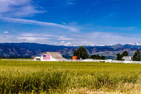 White barn with American flag painted on the side in front of growing wheat field near Rocky Mountains on summer dayのeditorial素材