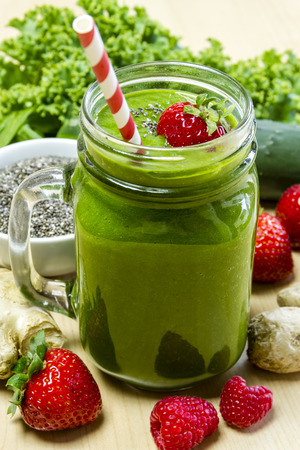 Healthy green juice smoothie surrounded by whole fruits, vegetables and chia seeds with fresh strawberry garnish and red swirl strawの写真素材