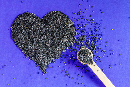 Black chia seeds arranged in heart shape with small spoon on purple background