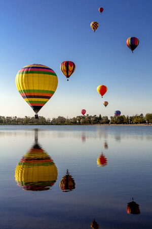 Multi-colored hot air balloons with bright blue sky flying over lake with reflectionの写真素材
