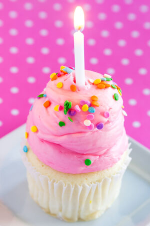 Vanilla cupcake with pink frosting sitting on plate with lit candle and pink polka backgroundの写真素材