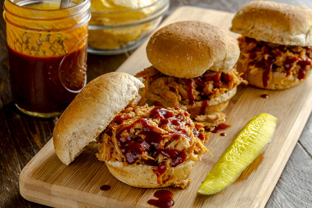 Three pulled pork barbeque sliders sitting on wooden cutting board with dill pickle with bottle of sauce and bowl of potato chipsの写真素材