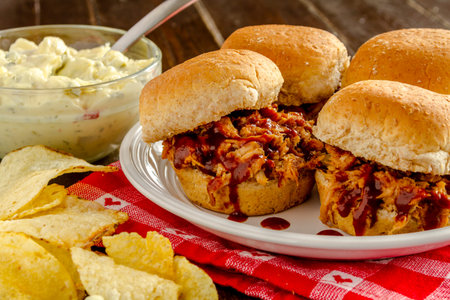 Four pulled pork barbeque sliders sitting on white plate with potato chips and potato salad on red...