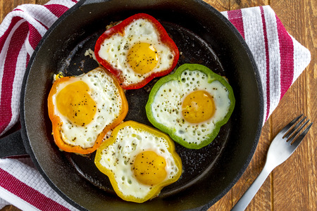 Close up of large cast iron skillet with fried eggs in green, yellow, red and orange bell peppers sitting on wooden table with fork and red striped towelの写真素材