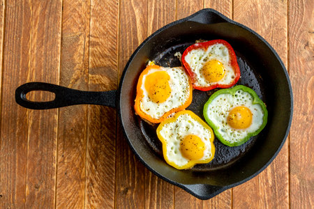 Large cast iron skillet with fried eggs in green, yellow, red and orange bell peppers sitting on wooden tableの写真素材