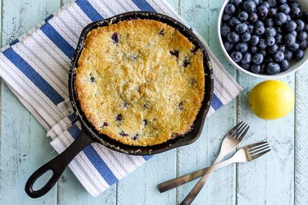 Homemade fresh blueberry cobbler baked in cast iron skillet pan with piece on white plate and with bowl of blueberries, lemon and forksの写真素材