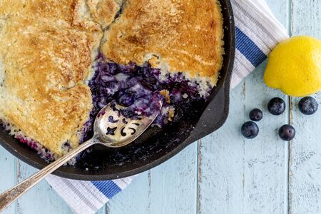 Close up of homemade fresh blueberry cobbler baked in cast iron skillet pan with large spoon and whole blueberries and lemonの写真素材
