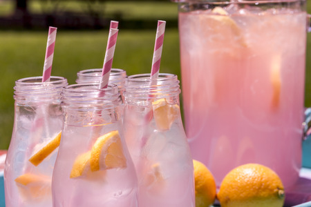 Close up of 4 small glass bottles and pitcher filled with fresh squeezed pink lemonade with pink swirled straws and lemon slices sitting on weather blue drink trayの写真素材