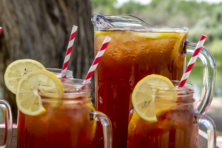 Pitcher and mason jar mugs filled with iced tea and lemons sitting on picnic table with red checked tableclothの写真素材