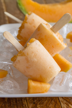 Homemade fresh pureed frozen cantaloupe melon popsicles on white plate with ice sitting on wooden table with fresh melonの写真素材