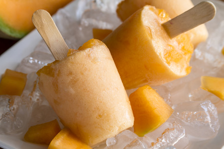 Close up of homemade fresh pureed frozen cantaloupe melon popsicles on white plate with ice sitting on wooden table with fresh melonの写真素材