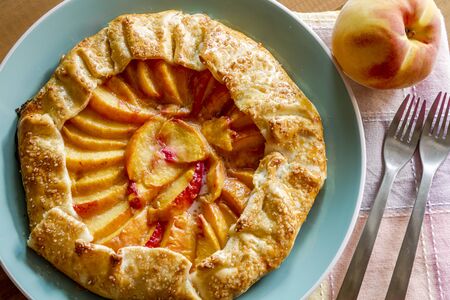 Homemade peach galette made with fresh organic peaches on blue plate with 2 folks and colorful napkinの写真素材