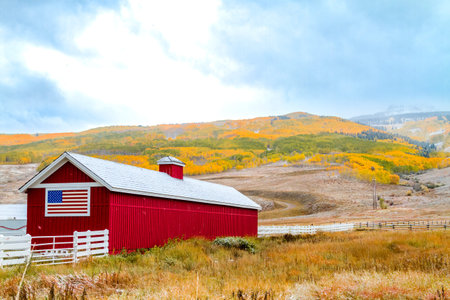 Red barn with American flag painted on side with mountainside filled with trees in full fall color with snow flurries and low clouds on mountainsのeditorial素材