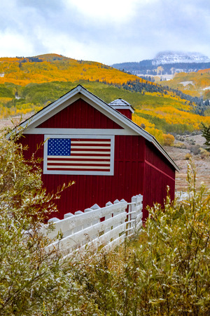 Red barn with American flag painted on side with mountainside filled with trees in full fall color and snow with low clouds on mountainsのeditorial素材
