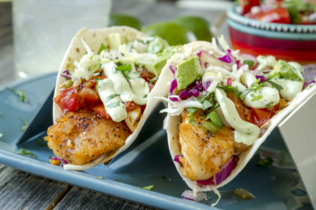 Close up of two white fish tacos with fresh coleslaw, salsa, lime cream and avocado sitting on blue plateの写真素材