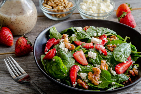 Gourmet organic strawberry spinach salad with feta cheese, walnuts and poppy seed dressing sitting in black bowl surrounded by salad ingredientsの写真素材
