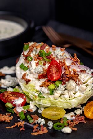 Classic restaurant ice berg wedge lettuce salad drizzled with blue cheese dressing, bacon, tomatoes, onions, chives sitting on black plates with dressing on the side. Vertical. Keto.の写真素材