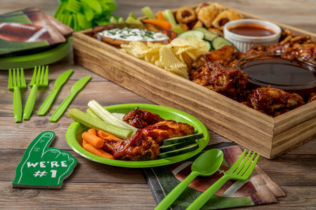 Football tailgate themed party food display with chicken wings, sauces, dips, wieners, vegetables, chips, pretzels in wooden tray with football themed plates, napkins and decorations shot sitting on wooden tableの写真素材